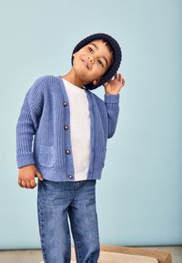 Cardigan tricotat albastru cu textură cu nervuri, închidere cu cinci butoane, două buzunare frontale, asociat cu un tricou alb și blugi albaștri, expus pe un fundal deschis.