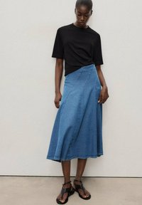 Chemise noire à manches courtes, jupe midi en denim bleu clair avec un design en A, et sandales plates noires. Détails de couture remarquables sur la jupe.