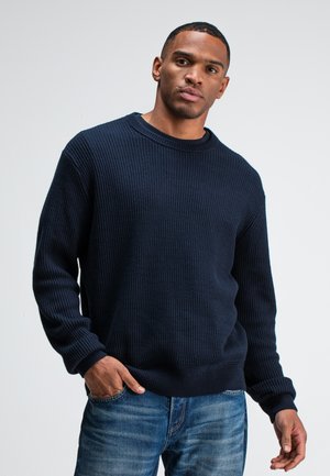 Navyblauwe geribde gebreide trui met lange mouwen en een ronde hals. De textuur is dik en comfortabel, gecombineerd met blauwe spijkerbroeken.
