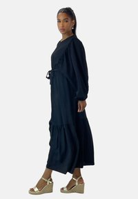 Vestido largo de manga larga en negro con volantes, corte relajado, cinturón para ajustar la cintura y tela con textura. Combinado con sandalias de cuña doradas.