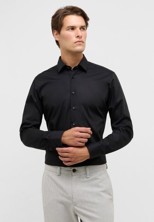 Camicia nera con bottoni, colletto e maniche lunghe. Tessuto liscio con una leggera lucentezza. Abbinata a pantaloni chiari con motivo.