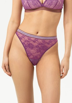 Femme portant une culotte taille haute en dentelle violette transparente et un bralette en dentelle assorti avec une ceinture rayée sur un fond uni.