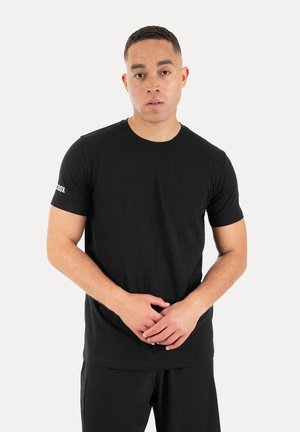 Mann steht mit verschränkten Händen, trägt ein schlichtes schwarzes T-Shirt mit kurzen Ärmeln und schwarze Hose vor einem einfarbigen hellen Hintergrund.