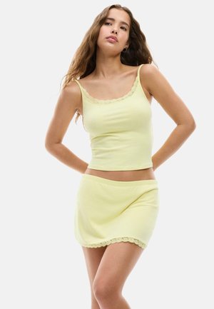 Jeune femme aux longs cheveux portant un top sans manches jaune pâle et une jupe assortie avec une bordure en dentelle, debout, les mains dans le dos.