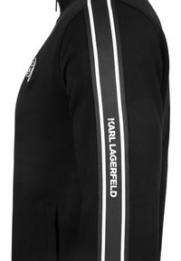 Sudadera negra con diseño de cremallera, bolsillos laterales y un detalle a rayas blancas y grises a lo largo de la manga izquierda con la marca "Karl Lagerfeld".