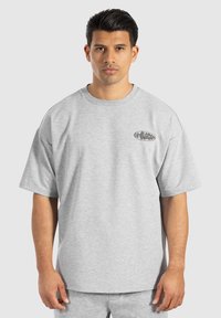 Camiseta gris extragrande hecha de una tela suave, con mangas cortas y un escote redondeado. En el frente presenta un pequeño detalle de logo negro.