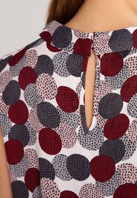 Cuello y parte superior de una blusa sin mangas con lunares rojos, grises y rosas, que presenta patrones moteados en negro y un escote tipo ojo de cerradura.