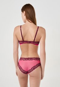 Donna con lunghi capelli castani, che indossa un set di reggiseno e slip in pizzo rosa e borgogna abbinati, in piedi contro uno sfondo chiaro e uniforme, di spalle.