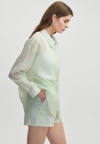 Camisa de color verde claro, de botones y de manga larga, con una tela texturizada y patrones sutiles y brillantes. El modelo posa con una mano en el bolsillo, vistiendo pantalones cortos.