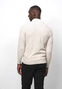 Maglione beige a maglia con maniche lunghe, texture a coste e silhouette aderente. Indossato con pantaloni neri, visto da dietro su uno sfondo bianco.