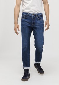 Jeans de mezclilla azul oscuro con corte recto, que cuentan con cinco bolsillos, costuras visibles y puños doblados. Combinados con zapatillas negras de caña alta.