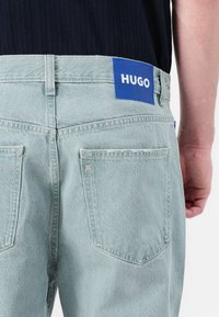 Világoskék farmernadrág, egyenes szabású, hátul zsebekkel, és egy kék HUGO címkével a deréknál. Felülete sima, anyaga puha.