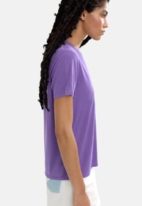 Fioletowa koszulka z krótkim rękawem, okrągłym dekoltem, wykonana z miękkiego materiału, o prostym kroju i gładkiej fakturze. Prosty design.