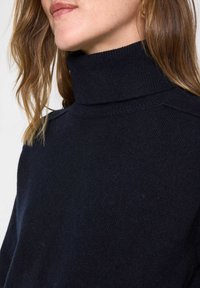 Donna che indossa un maglione a collo alto a coste di un blu navy scuro, con dettagli sulle cuciture delle spalle e capelli ondulati di un marrone chiaro.