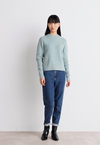 Hellblauer gerippter Pullover mit rundem Ausschnitt, kombiniert mit dunkelblauen Jeans, die an den Bündchen leicht hochgekrempelt sind, und schwarzen Knöchelschuhen.