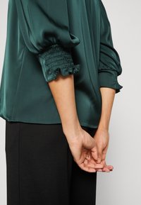 Blouse en satin vert émeraude avec des manches ballons, poignets élastiqués ornés de bords froncés ; associée à un pantalon noir ajusté.