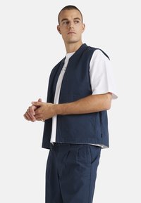 Chaleco sin mangas azul marino de tela de algodón, con un bolsillo frontal y un corte relajado, usado sobre una camiseta blanca y combinado con prendas de abajo a juego.