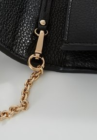 Coin de sac en cuir noir texturé avec un fermoir en métal doré attachant une bandoulière chaîne dorée, présenté sur un fond gris clair.