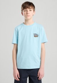 T-shirt in cotone azzurro chiaro con una grafica di un skateboarder ricamata sul lato sinistro del petto. Maniche corte, design a girocollo.