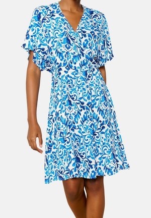 Femme portant une robe blanche mi-longue avec des motifs floraux et tourbillonnants bleus et des manches courtes évasées, debout avec un bras détendu.