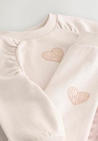 Világos rózsaszín pulóver fodros vállvarrással és dombornyomott szívmintákkal az elején és az ujján. Puha pamut anyagból készült.