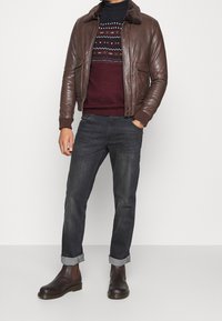 Mann in brauner Lederjacke, gemustertem Pullover, dunklen, umgeschlagenen Jeans und dunkelbraunen Chelsea-Stiefeln, der vor einem schlichten Hintergrund steht.