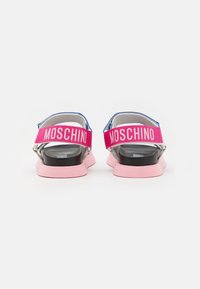 Takana näkymä vaaleanpunaisista ja mustista sandaaleista, joissa on paksut vaaleanpunaiset remmit ja joissa on "MOSCHINO"-teksti valkoisella taustalla.