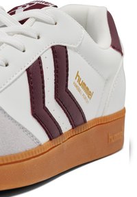 Witte sneaker met bordeaux accenten en suède details. Kenmerken zijn een gouden logo, veterontwerp en een gestructureerde gumrubberen zool. Klassieke silhouet.