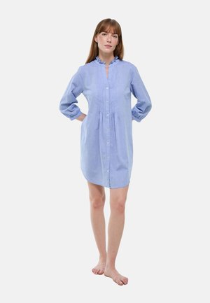 Femme debout pieds nus portant une robe bleu clair à manches longues, boutonnée avec un col à volants sur fond blanc.