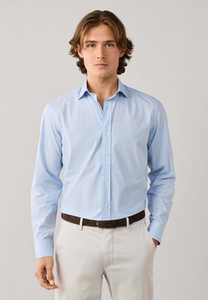 Joven con el pelo castaño de longitud media lleva una camisa azul claro, pantalones beige y cinturón marrón, de pie con las manos en los bolsillos.