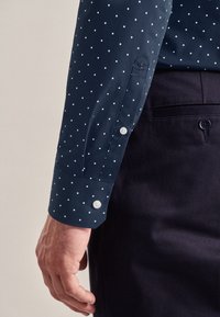 Donkerblauw shirt met witte stippen, lange mouwen, knoopmanchetten en subtiele borduurdetails op het mouwsplit. Draag met een donkere broek.