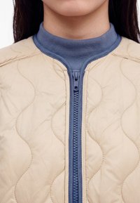 Primer plano de una persona que lleva una chaqueta beige acolchada con una cremallera azul y un cuello alto acanalado azul debajo.