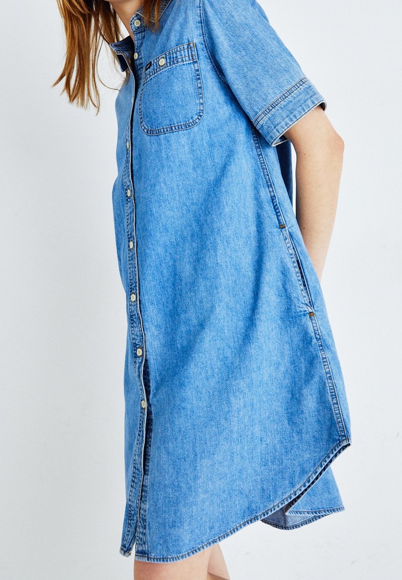Personne portant une robe-chemise en jean bleu clair à manches courtes, avec des boutons et une poche poitrine, debout devant un fond blanc uni.