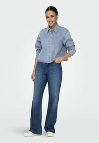 Denim jacket in light blue with a collar, button closure, and long sleeves; paired with wide-leg dark blue jeans and white sneakers.