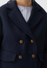 Marinblå dubbelknäppt kappa i strukturerad tyg med guldfärgade knappar och två framfickor, med en bred krage.