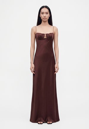 Jeune femme aux longs cheveux raides porte une robe en satin marron longue jusqu'au sol avec des bretelles fines et un corsage sculpté, debout sur un fond blanc.