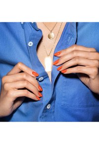 Camicia blu con bottoni e taschino sul petto; mani con smalto arancione brillante che tengono una collana in oro con un pendente circolare e un pendente rettangolare.
