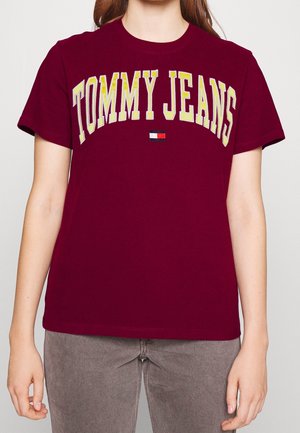 Osoba ubrana w bordową koszulkę z krótkim rękawem z żółtym napisem "TOMMY JEANS" oraz małym logo w postaci czerwono-biało-niebieskiej flagi na piersi.