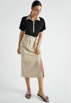 Vrouw draagt een zwart poloshirt met beige kraag en knopen, een beige midi-rok met zijschuif en strik, en sandalen met luipaardprint en strik.