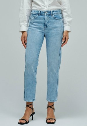 Mujer con vaqueros azul claro de talle alto y pierna recta, camisa blanca abotonada metida por dentro y sandalias negras de tacón con tiras.