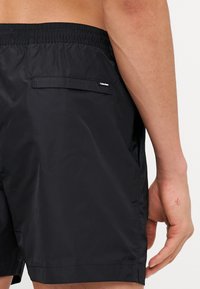 Svart badshorts laget av lett, vannavstøtende materiale. Har elastisk midje og en baklomme med etikett.