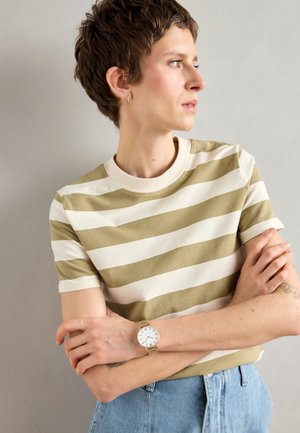 T-shirt rayé en vert olive et blanc, à manches courtes, avec un col côtelé. Le modèle porte une montre en mailles argentées ; des jeans en denim sont également visibles.