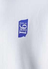 Camiseta de tela blanca con un logo rectangular morado que lleva el texto "REPLAY 69 cero". El diseño incluye líneas limpias y un formato sencillo.