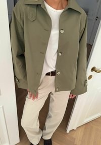 Osoba oblečená v olivovo zelenej zapínanej bunde, bielej košeli, béžových nohaviciach a čiernych topánkach, stojaca v dverách na drevenej podlahe.