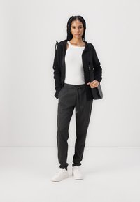 Haut zippé noir, débardeur blanc, pantalon de survêtement gris avec cordon de serrage, et baskets blanches. Modèle tenant un sac noir, se tenant sur un fond neutre.