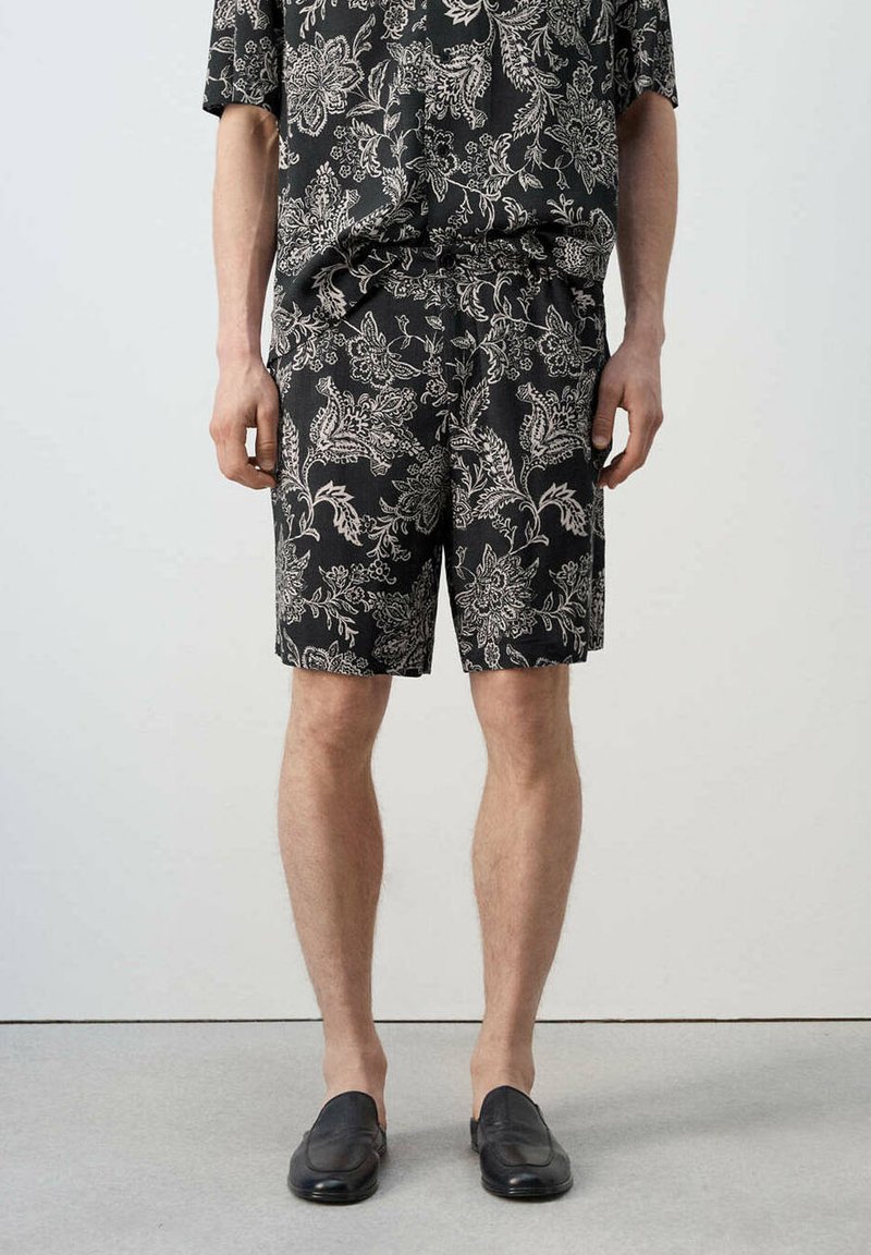Homme portant un short noir à motif floral et une chemise assortie avec des chaussures noires slip-on, debout devant un fond uni.