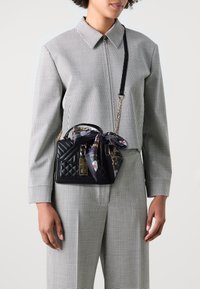 Femme en costume à carreaux noir et blanc portant un sac à main matelassé noir avec une chaîne dorée et une écharpe florale nouée sur la poignée.