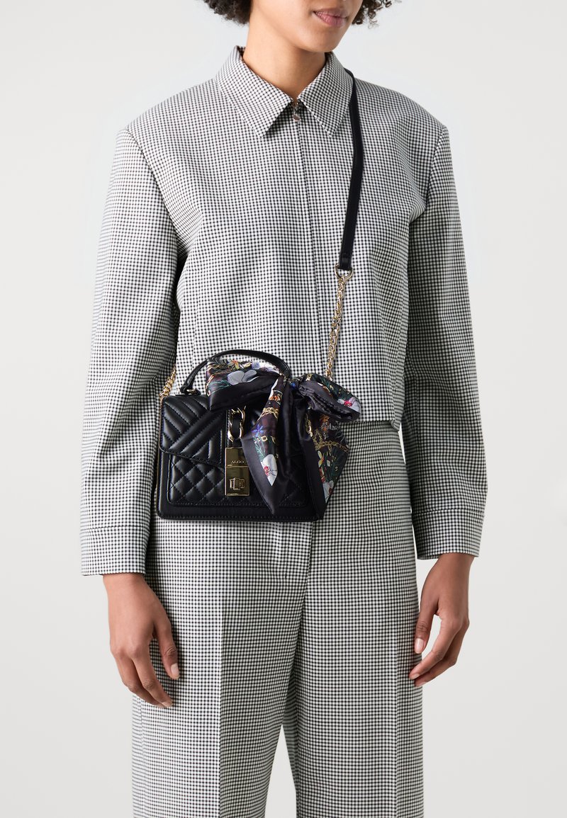 Femme en costume à carreaux noir et blanc portant un sac à main matelassé noir avec une chaîne dorée et une écharpe florale nouée sur la poignée.