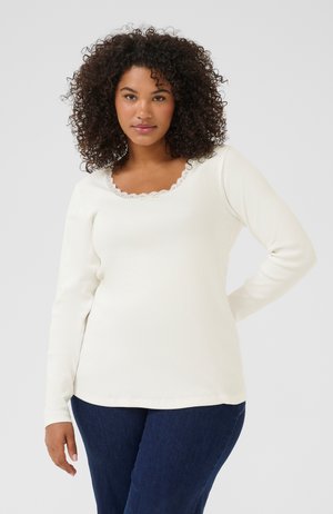 Langsleeve crèmekleurige top met een geribbelde structuur en golvende kanten randen rond de ronde halslijn. Gecombineerd met donkerblauwe spijkerbroek.