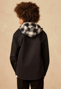 Schwarze Jacke mit kariertem Kapuzenfutter, das eine glatte Textur und eine lässige Passform aufweist, von hinten betrachtet.